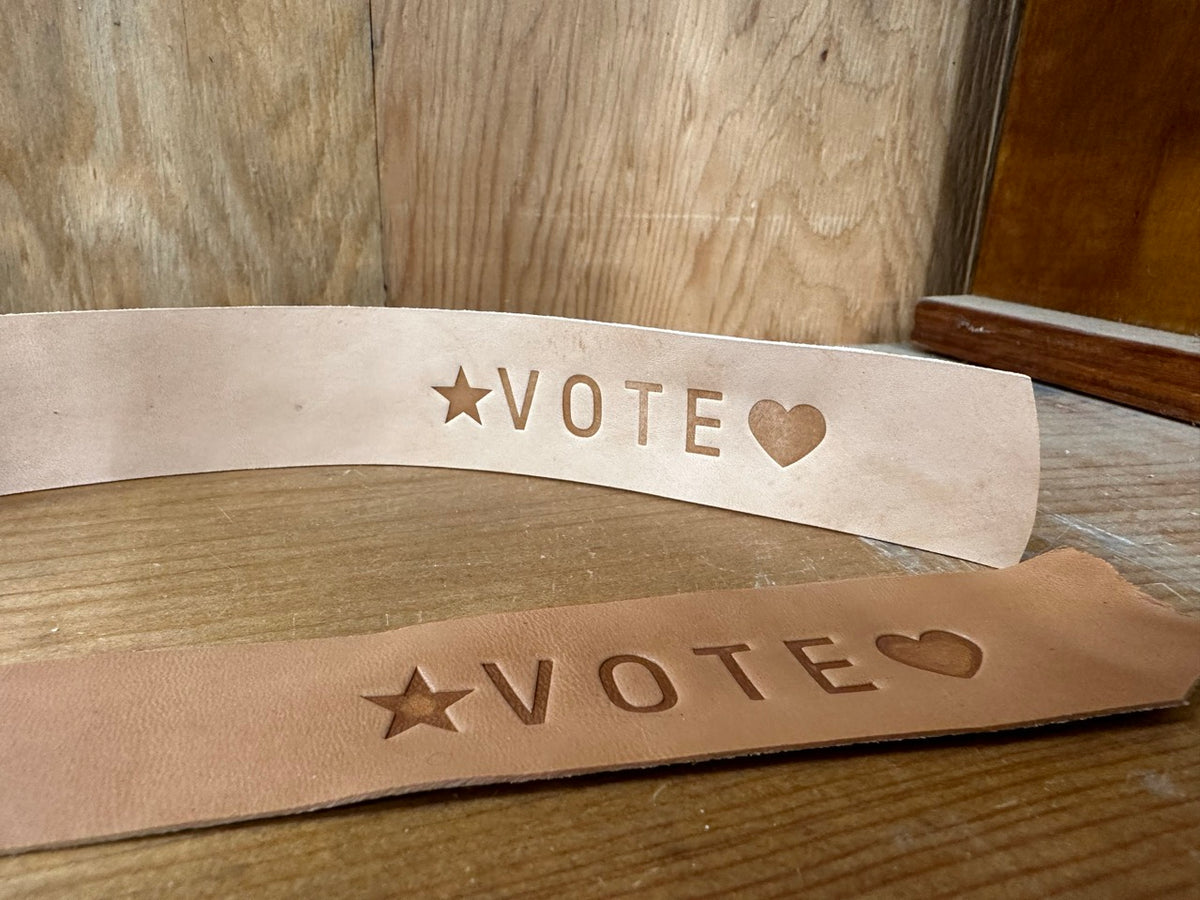 Two natural leather straps with the word VOTE imprinted on it with a star and a heart propped up on a wood shelf to display