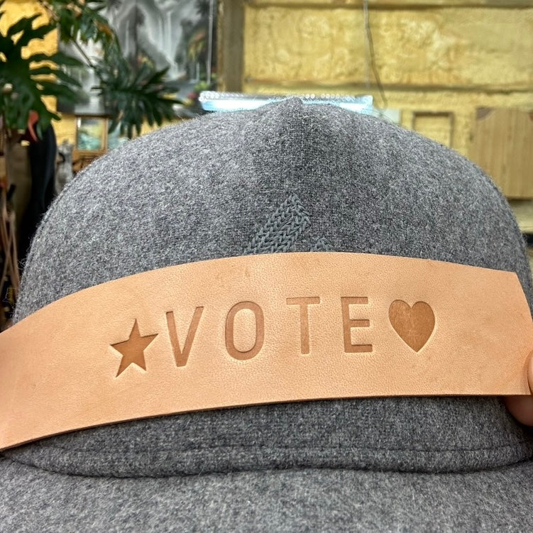 A natural leather strap with the word VOTE imprinted on it with a star and a heart held up against a baseball cap