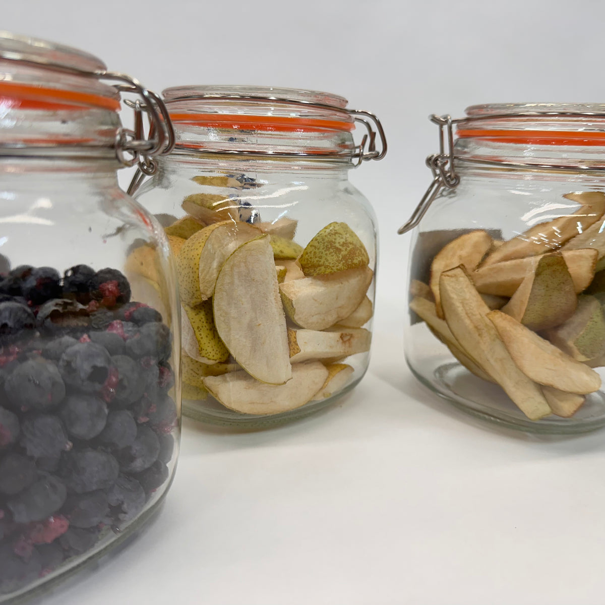 Three glass jars with cage tops containing different freeze-dried fruits on a white background: blueberries, Asian pears, and pears
