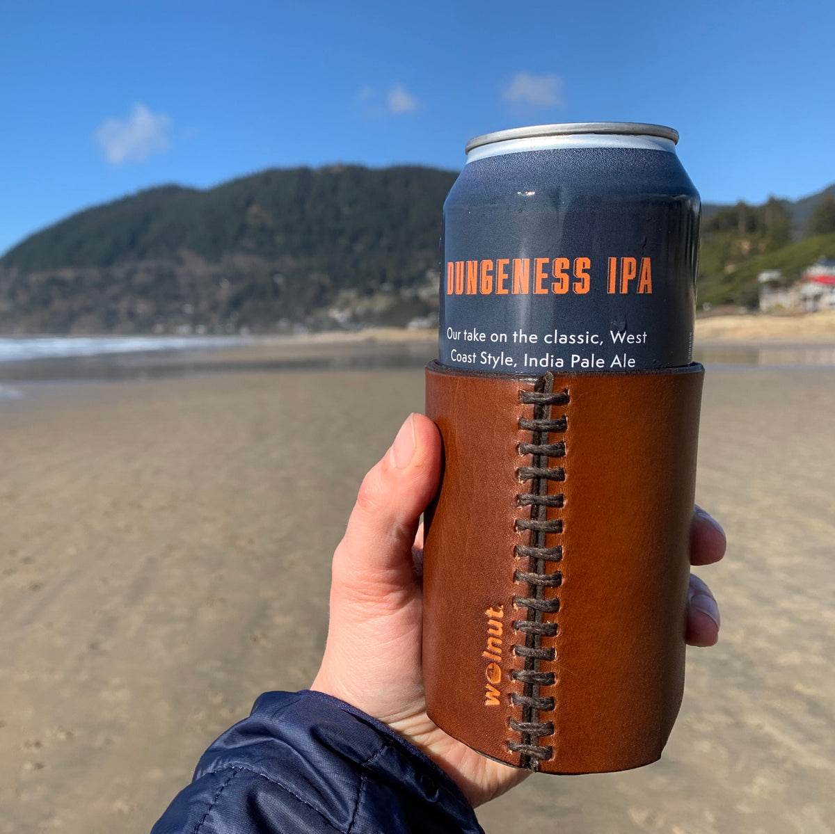 Hand holding a tallboy can of Dungeness IPA beer with a hand-stitched leather can sleeve on a beach with a mountain in the background on a sunny day
