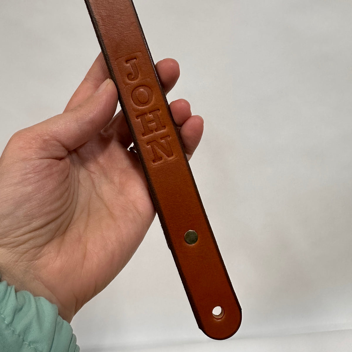 Hand holding a Tilikum leather handle with 'JOHN' embossed on it (reading vertically) against a white background