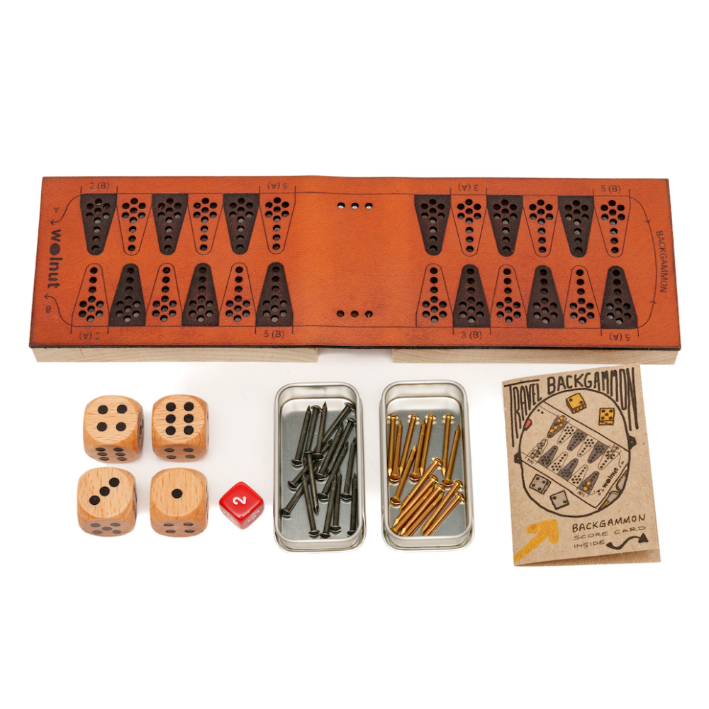 White background photo of a leather folding travel cribbage board opened up to full size and displaying all the parts beside it: dice, doubling cube, brass and black game pieces (which are small nails) and a paper booklet that says Travel Backgammon, Score Card Inside