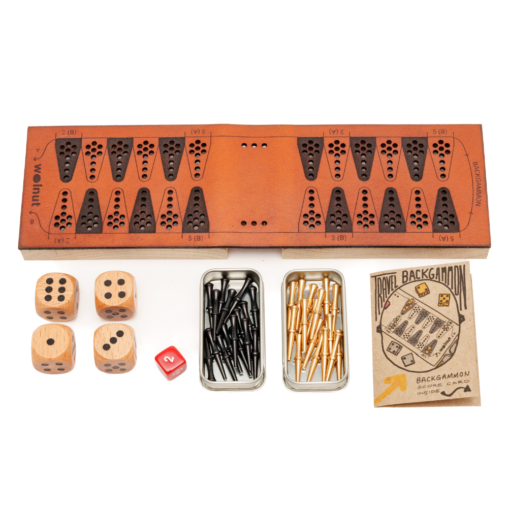 White background photo of a leather travel backgammon board opened up and all the parts to the side: wood dice, a small red doubling cube, slider tins with brass and black pegs, and a little booklet with score card