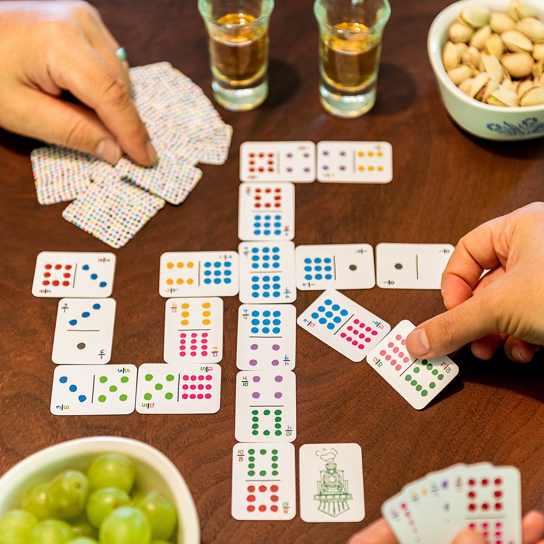 Two people playing dominoes using small, domino-sized playing cards on a walnut wood table with snacks and drinks. The domino cards are double 12 with different colors for each number and hand-drawn.