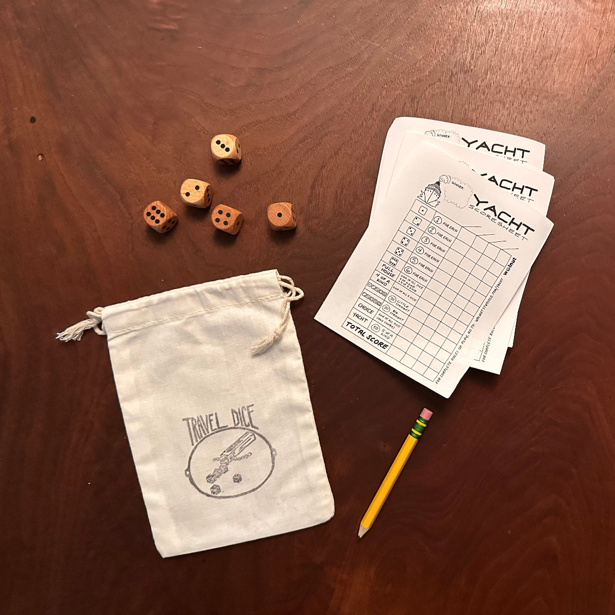 All parts of the Travel Yacht kit laid out on a walnut wood table: muslin carrying pouch stamped with a Travel Dice graphic, multiple sheets of Yacht (Yatzy) scoresheets, a sharp Ticonderoga golf pencil with an eraser, and five wood dice
