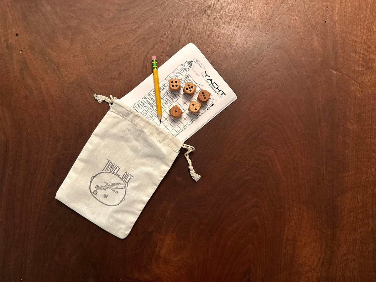 All parts of the Travel Yacht kit laid out on a walnut wood table, with the contents spilling out of the bag: muslin carrying pouch stamped with a Travel Dice graphic, multiple sheets of Yacht (Yatzy) scoresheets, a sharp Ticonderoga golf pencil with an eraser, and five wood dice
