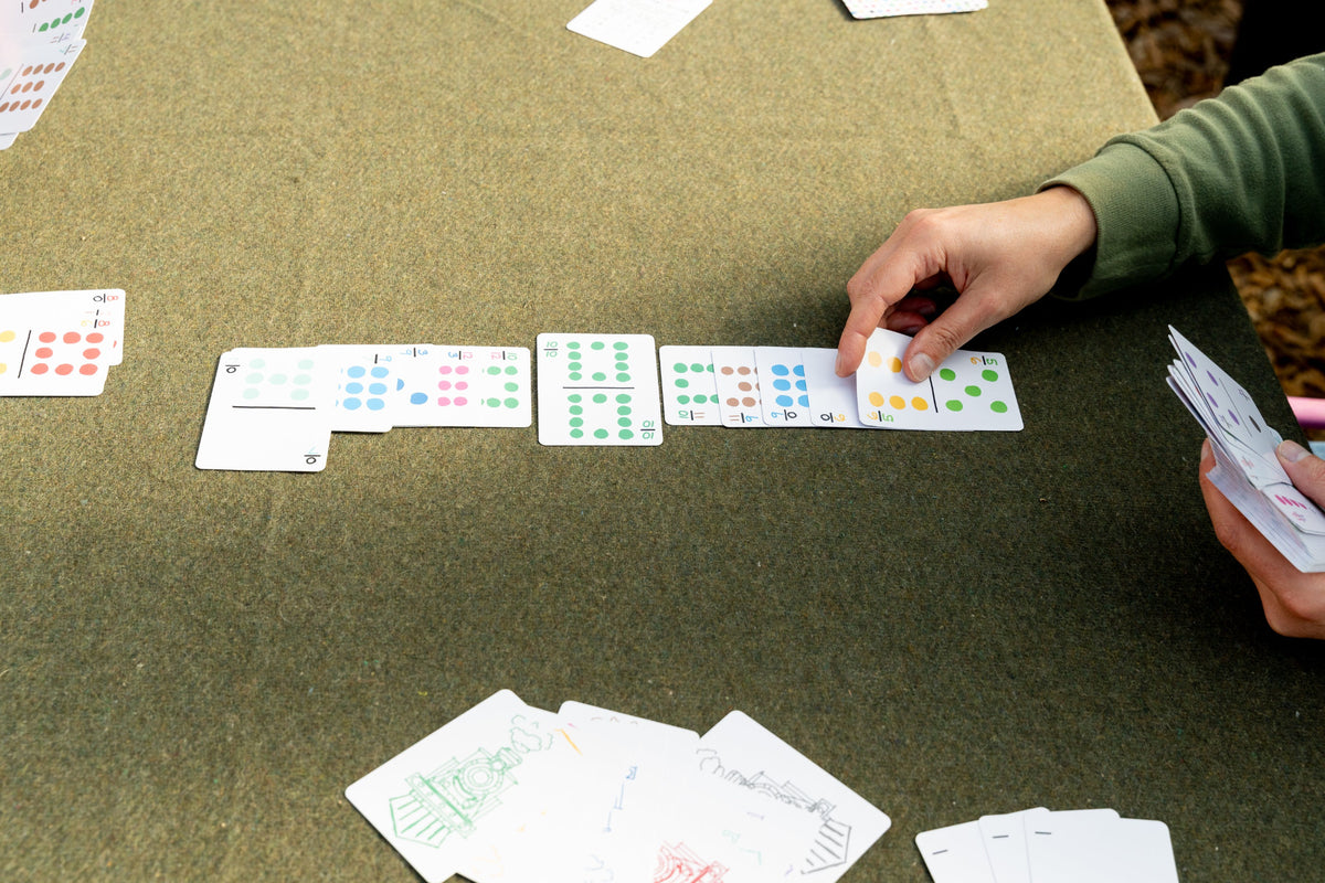 A close-up of a person's hands playing a card game on a green felt table, with cards showing colored dots and handwritten notes scattered nearby.