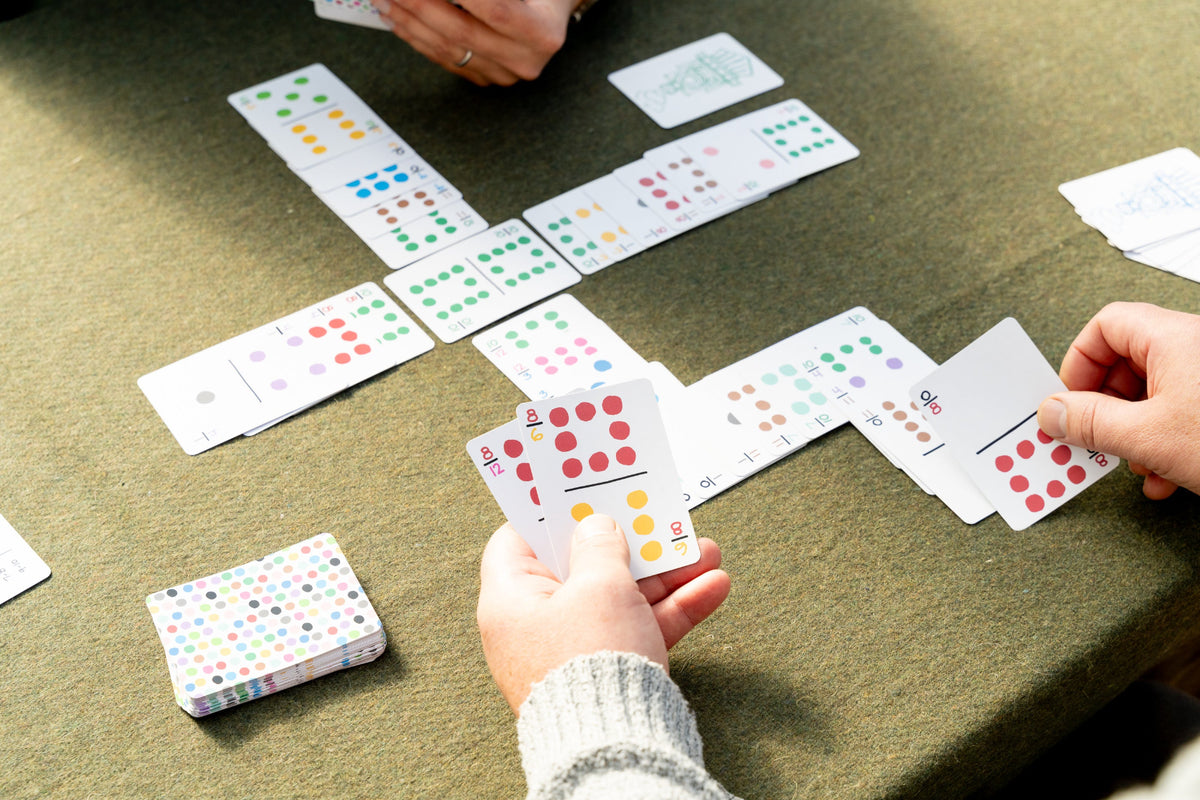 A close-up of a card game in progress on a green felt table, with several hands visible holding and placing patterned playing cards. A small deck box sits nearby.
