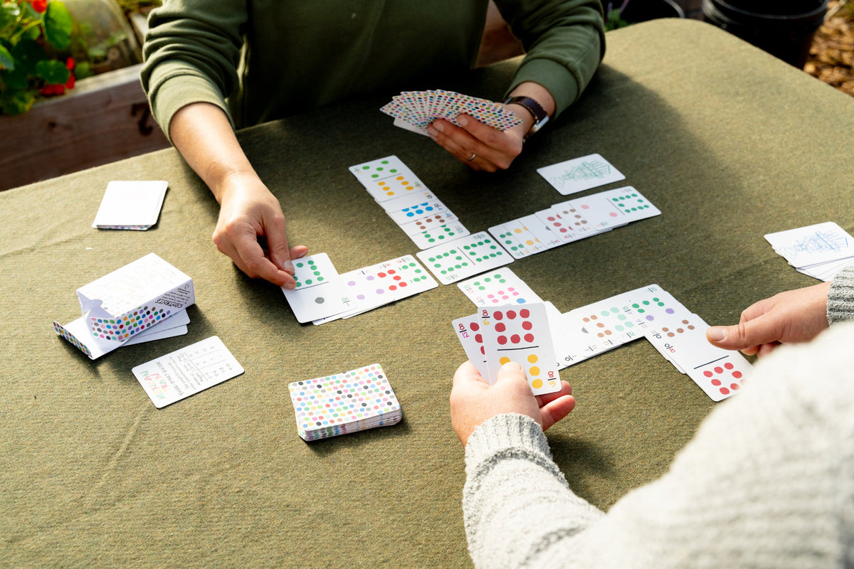 Two people playing a card game outdoors at a table covered with a green cloth. The cards are spread out in a cross shape, and several small stacks of cards are visible on the table.