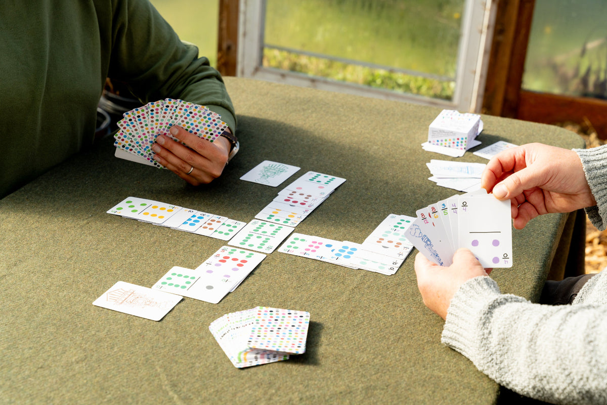 Two people sitting at a table playing with a deck of cards. One person on the left holds a hand of cards in a green sweater, and another person on the right holds cards in a gray sweater.
