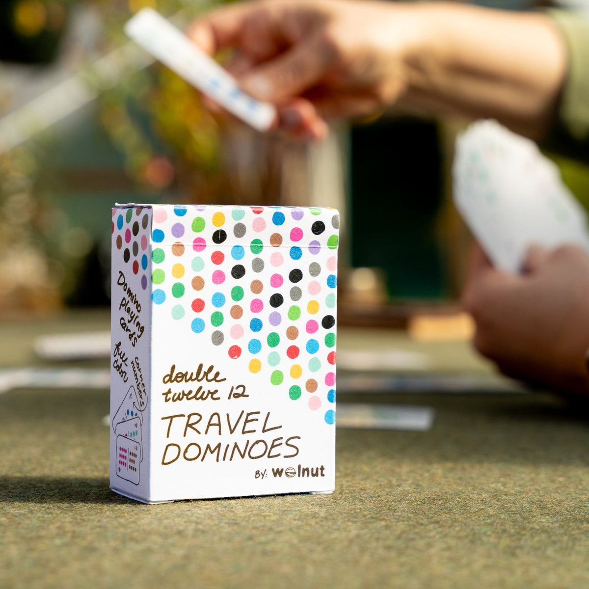 Close-up of a playing card size box of double 12 travel domino playing cards on a table while in the background people are playing cards blurred out