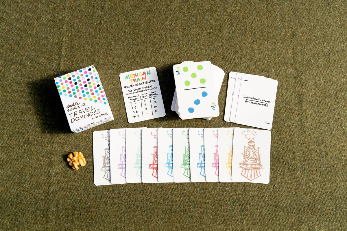 Train-themed domino card game set laid out on an olive green textured surface, including a colorful box, instruction card, playing cards, and a small item for scale.