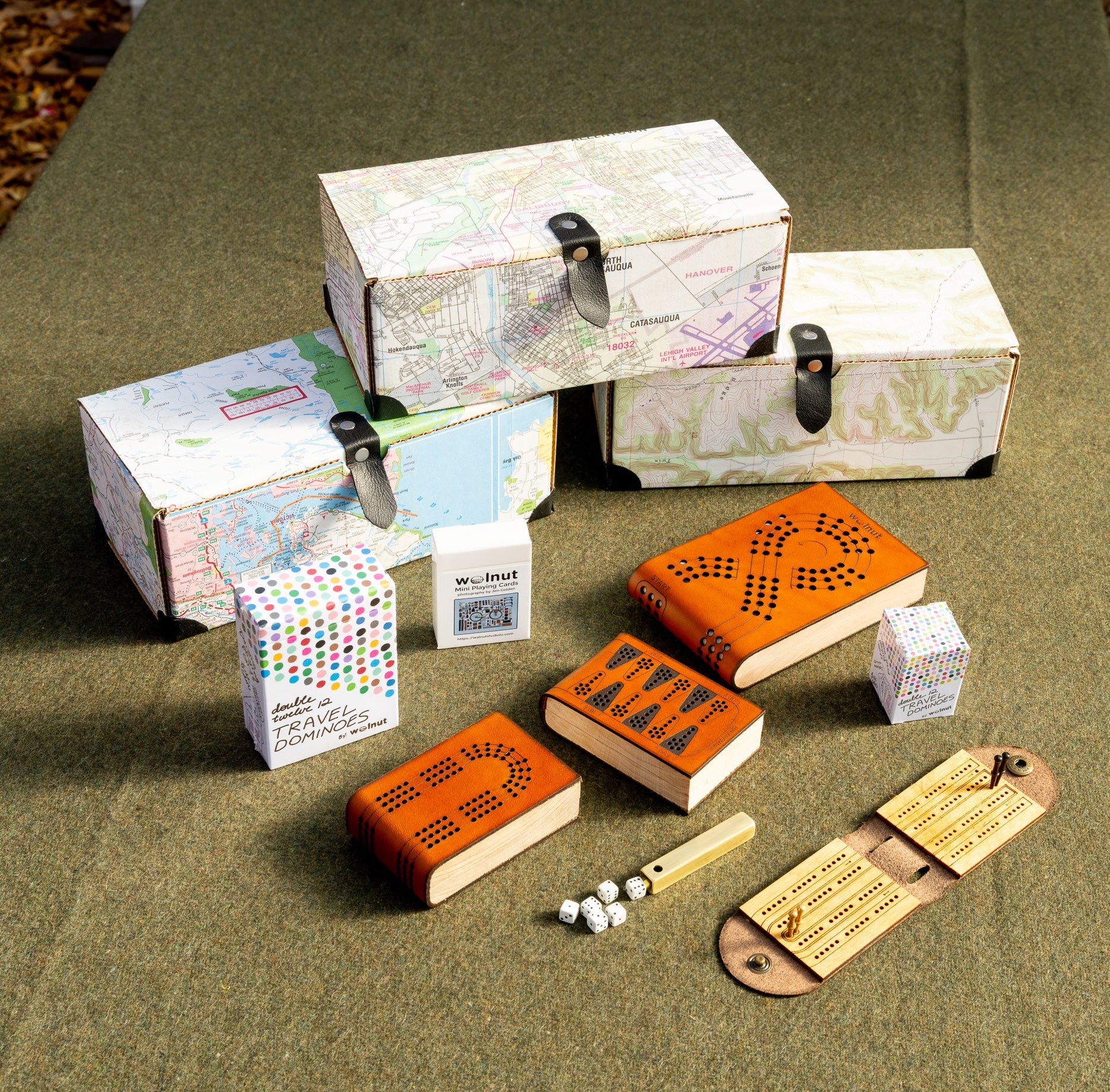 A group of map-covered gift boxes, and a display of Walnut Studiolo pocket-sized travel games on a green wool army blanket: backgammon, cribbage, dice, and dominoes