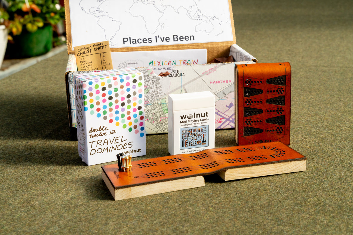 &quot;Game Lovers&quot; Travel games gift bundle shown with a handcrafted memory box, &quot;places I&#39;ve been&quot; keepsake map, printed scorecards for Mexican Train, and cribbage counting cheatsheet, leather cribbage board, half-size travel playing cards, leather backgammon board, and double 12 travel dominoes playing cards 