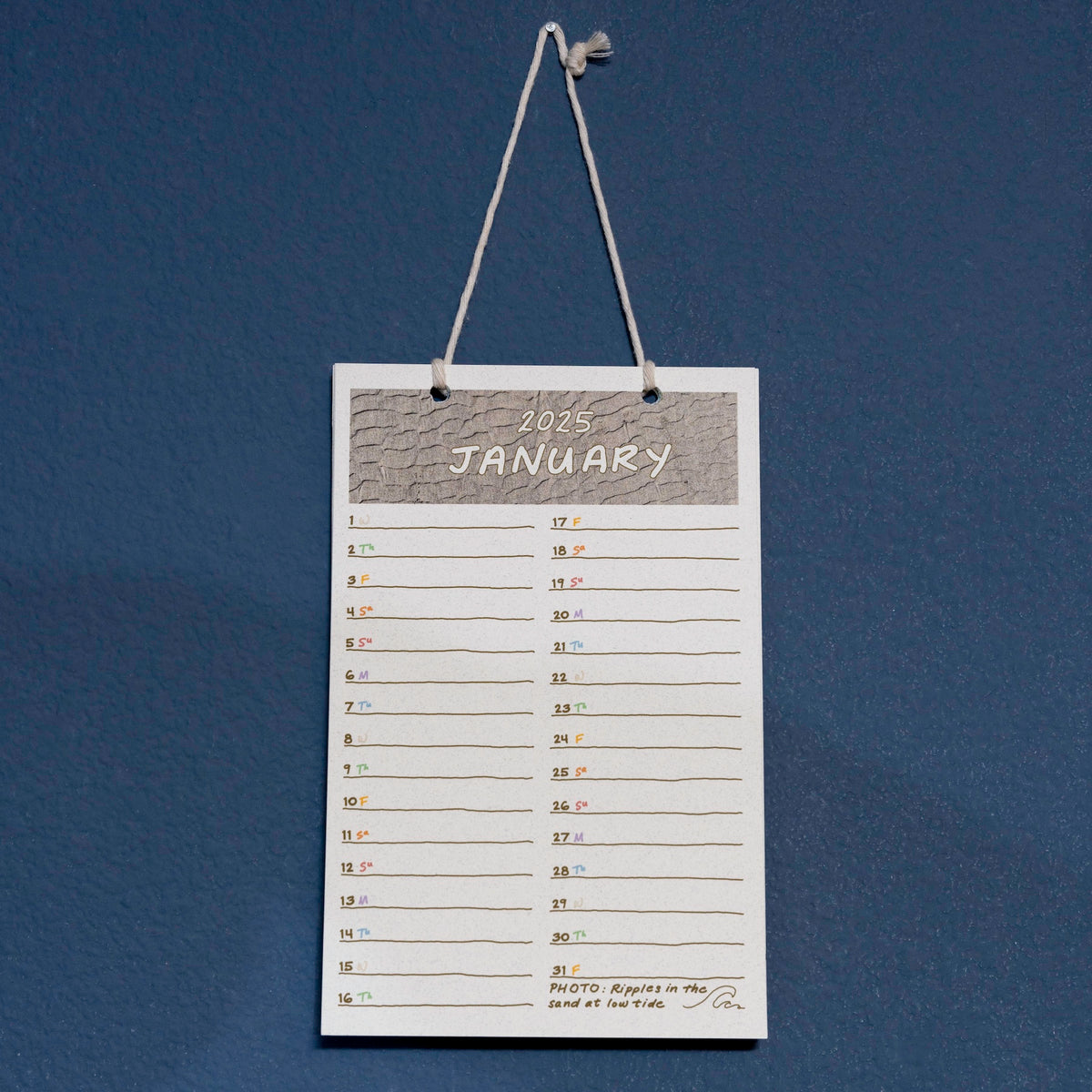 Calendar hanging on the wall showing the month of January, with a beach sand photo, a small drawing, and a line for each day for a short journal or diary note, with color-coded days of the week