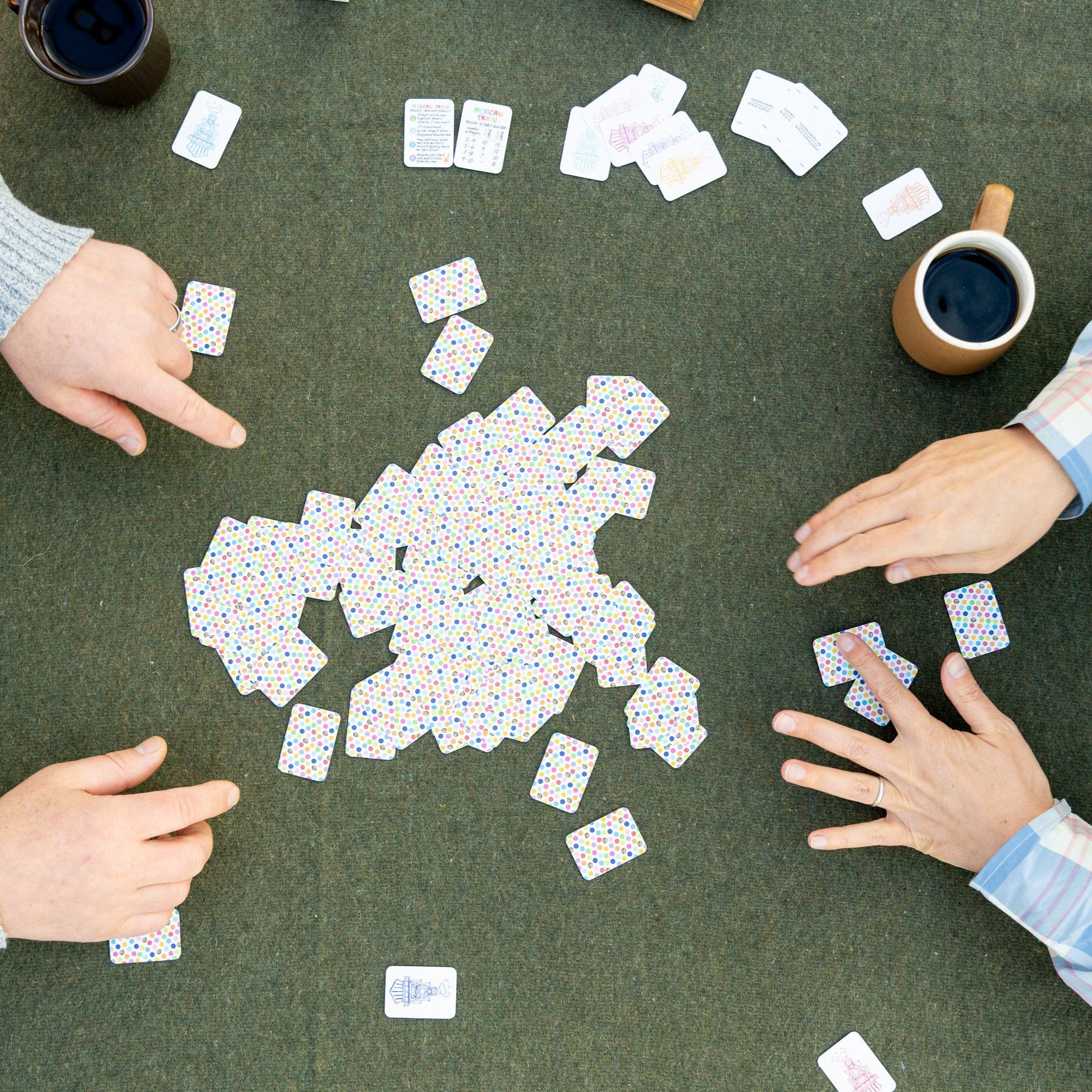 Bird's eye view of a table while two people are playing domino and picking their hands from the pile of tiles or boneyard, using the new version 2 of the double 12 travel domino playing cards. The table has a green wool background and they are drinking cups of coffee. 