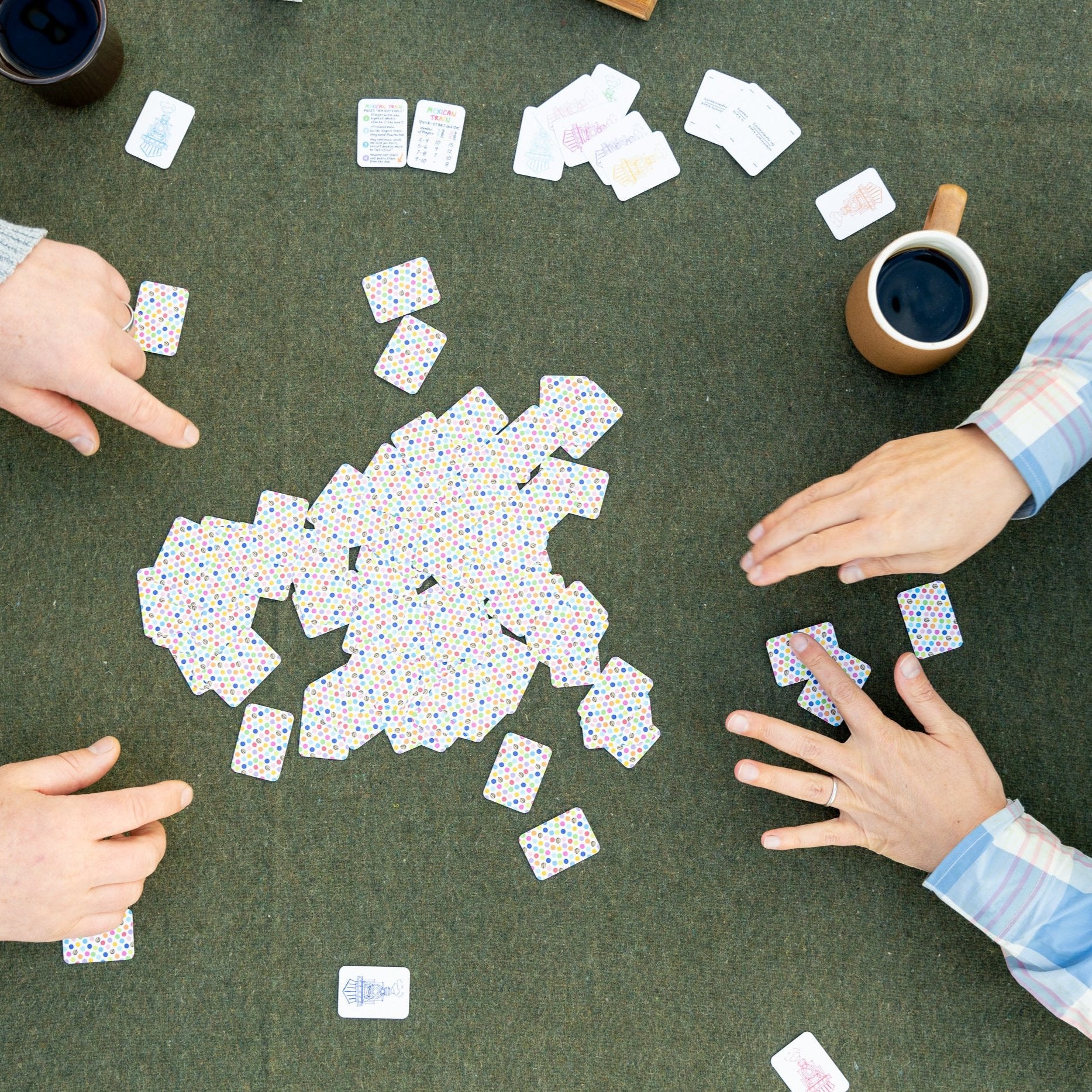 Bird's eye view of a table while two people are playing domino and picking their hands from the pile of tiles or boneyard, using the new version 2 of the double 12 travel domino playing cards. The table has a green wool background and they are drinking cups of coffee. 