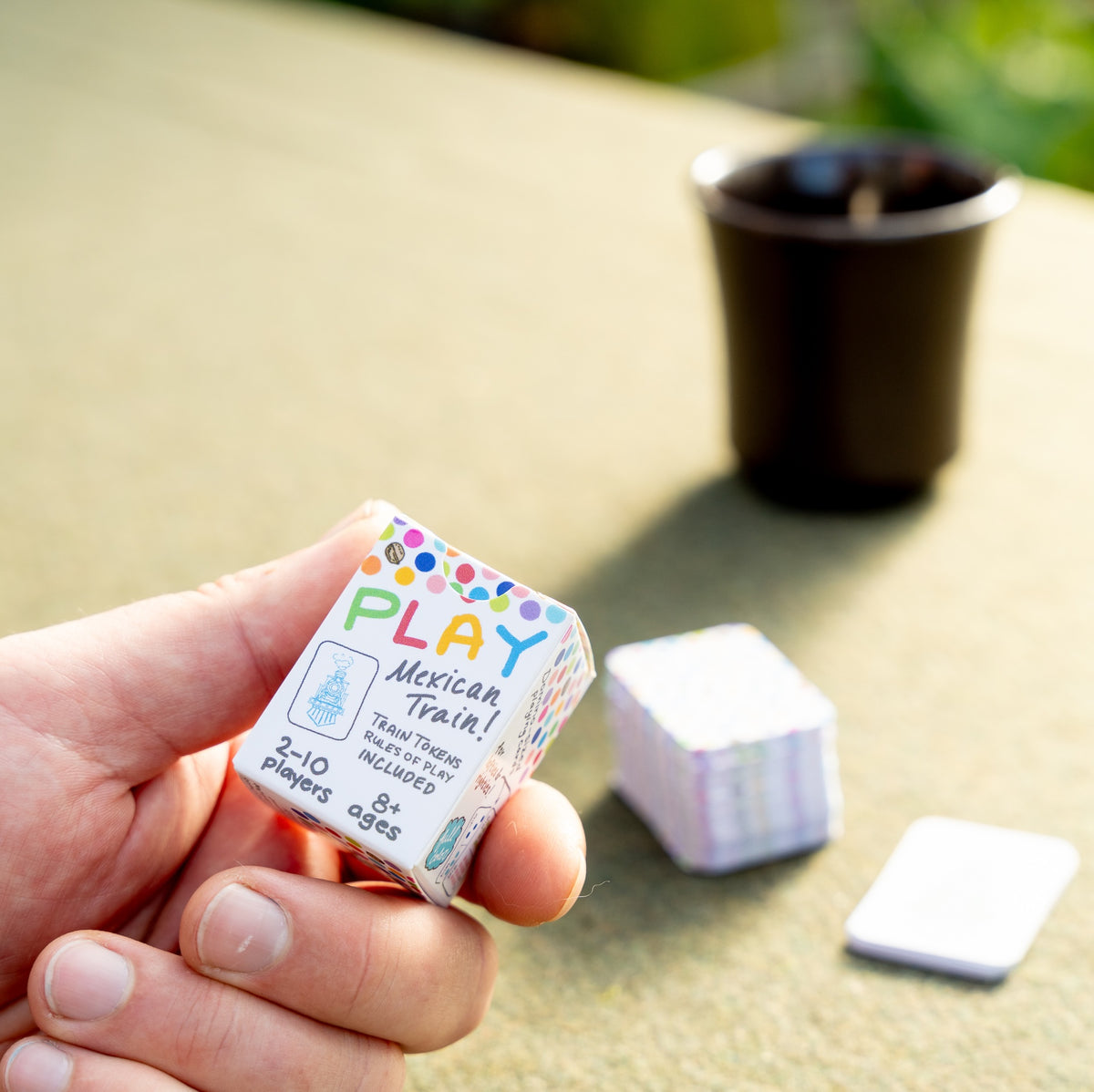 A man&#39;s hand holding the small box of travel domino playing cards between his thumb and forefinger. Blurred out in the background is a sunny table with a green tablecloth, the stack of cards ,and a cup of coffee. On the box, he&#39;s reading, Play Mexican Train! 2-10 Players, Ages 8+