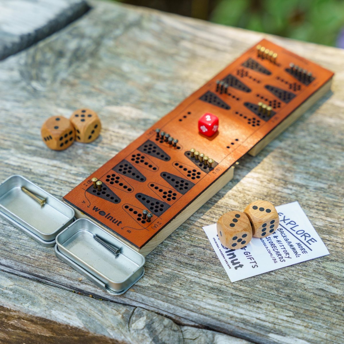 A travel backgammon board with matte, natural materials and accessories, wood dice and brass and black small nail game pieces, is on a table and catching the sunlight in a natural outdoor setting