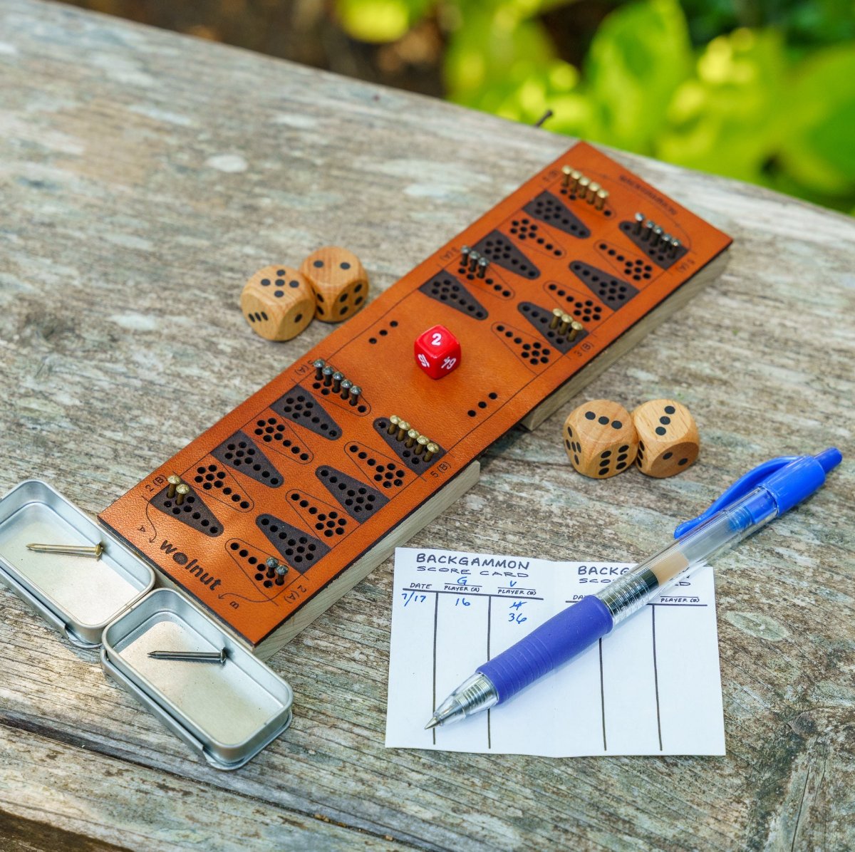 A travel backgammon board set up on a weathered wood picnic table, ready to play for two people and a scorecard with a pen for scale. 