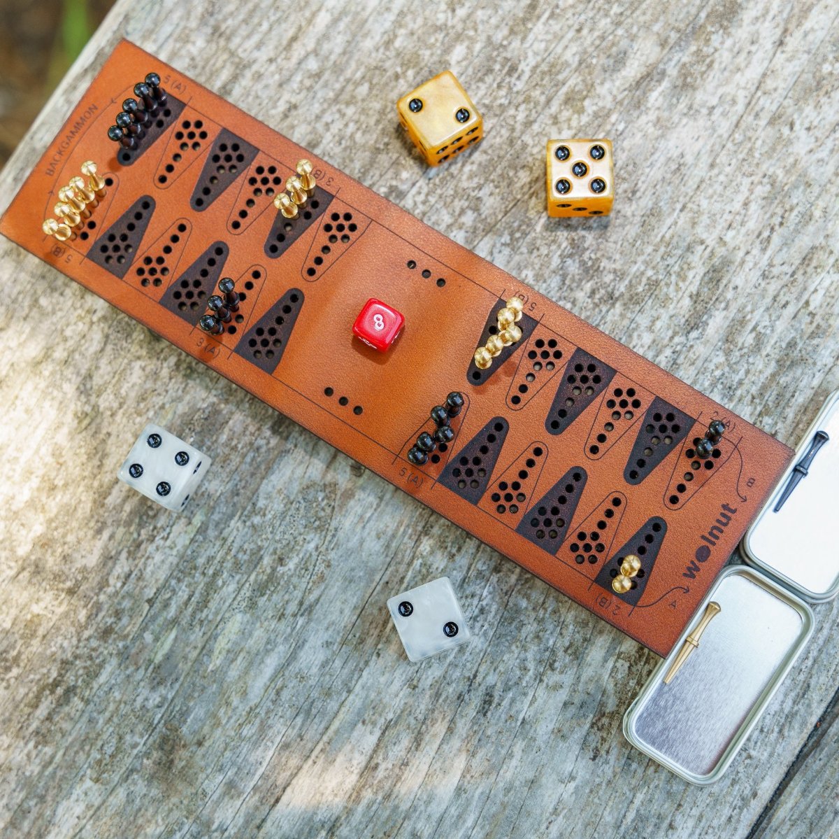 A travel backgammon board with shiny accessories, bright marbleized gold and gray dice and brass and black peg game pieces, is on a table and catching the sunlight in a natural outdoor setting