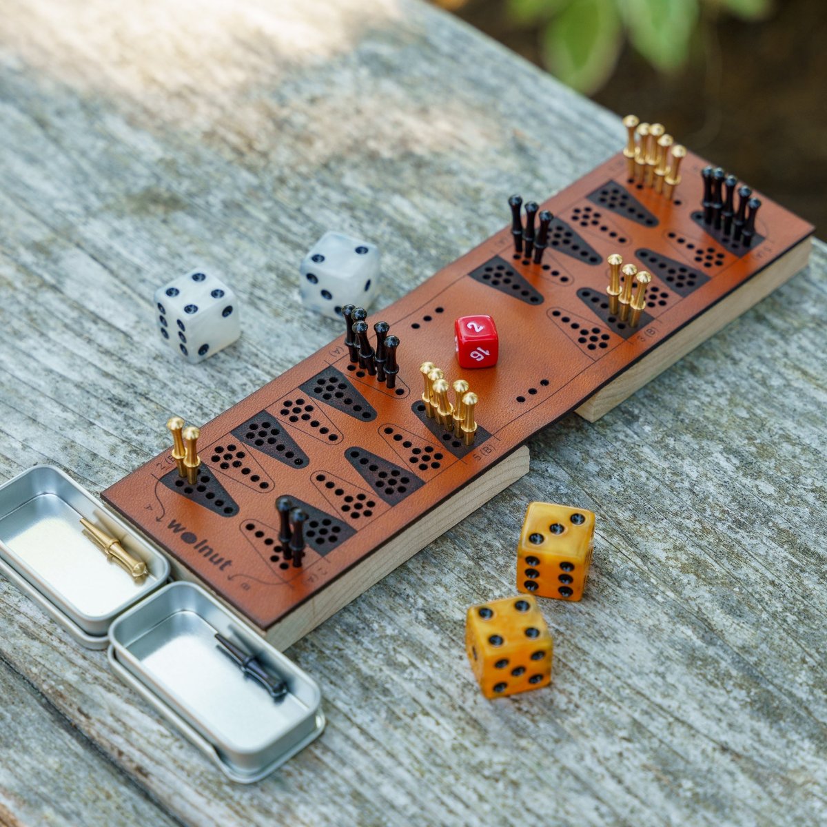 A travel backgammon board with shiny, eye-catching materials and accessories, luminous marbleized pearl and gold resin dice and brass and black small cribbage peg game pieces, is on a table and catching the sunlight in a natural outdoor setting