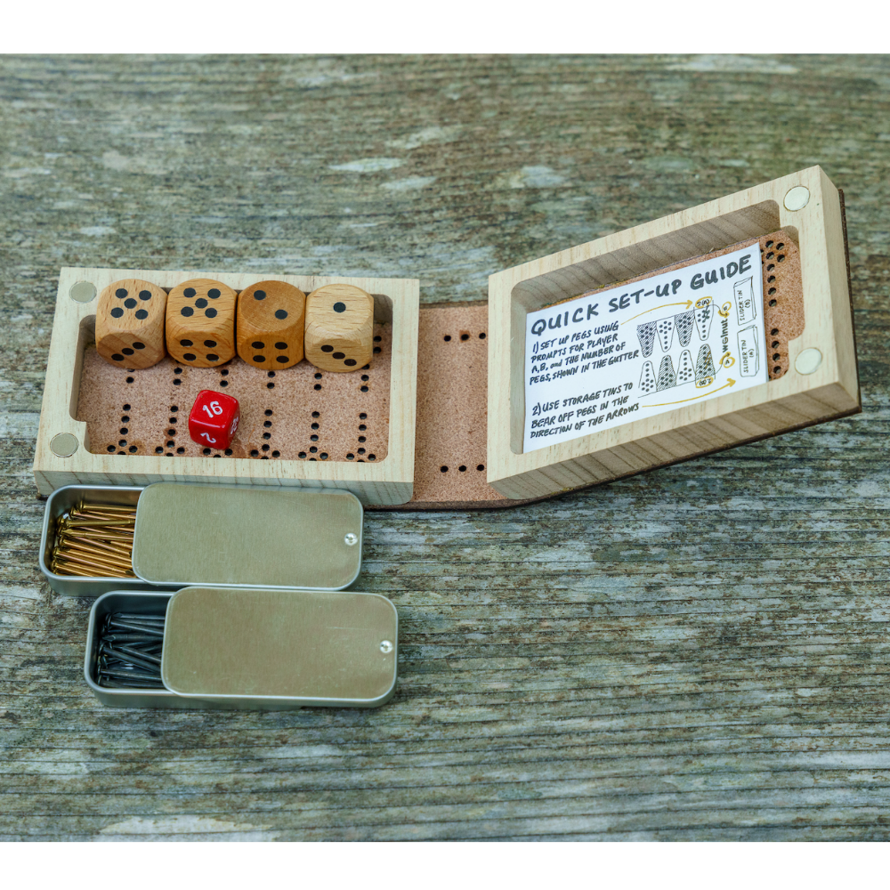 A leather travel backgammon board is lying on a weathered wood surface, opened up like a book so you can see the inside. Inside there are four dice, a doubling cube, and a small booklet neatly fit inside with empty space for two slider tins of pegs, which sit off to the side of the board with the top slid partially open to show the contents, which are brass and black game pieces