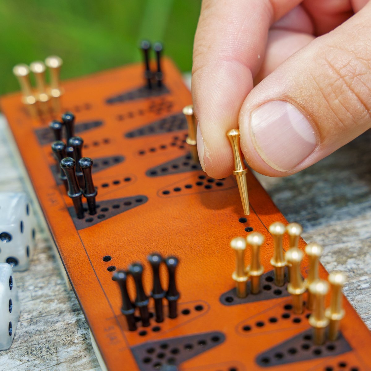 Close-up photo of a small leather travel-sized backgammon board that uses brass and black pegs instead of checkers on top of a weathered wood picnic table. A man&#39;s fingers can be seen picking up and moving a brass peg along the board