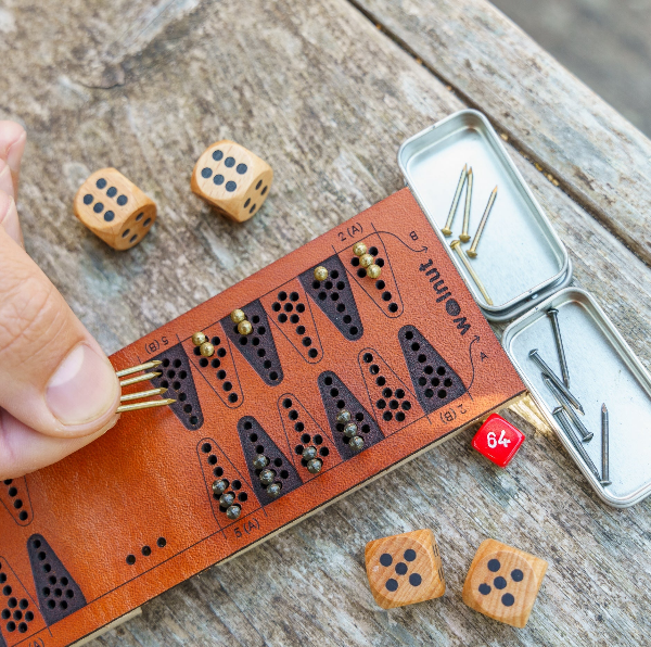 Close-up of a folding leather travel backgammon board mid-play on a weathered wood surface. A hand is reaching in and moving four game pieces (which are brass nails) between triangles, which are laser-engraved in the honey dyed leather. Accessories are visible, dice and doubling cube and peg storage