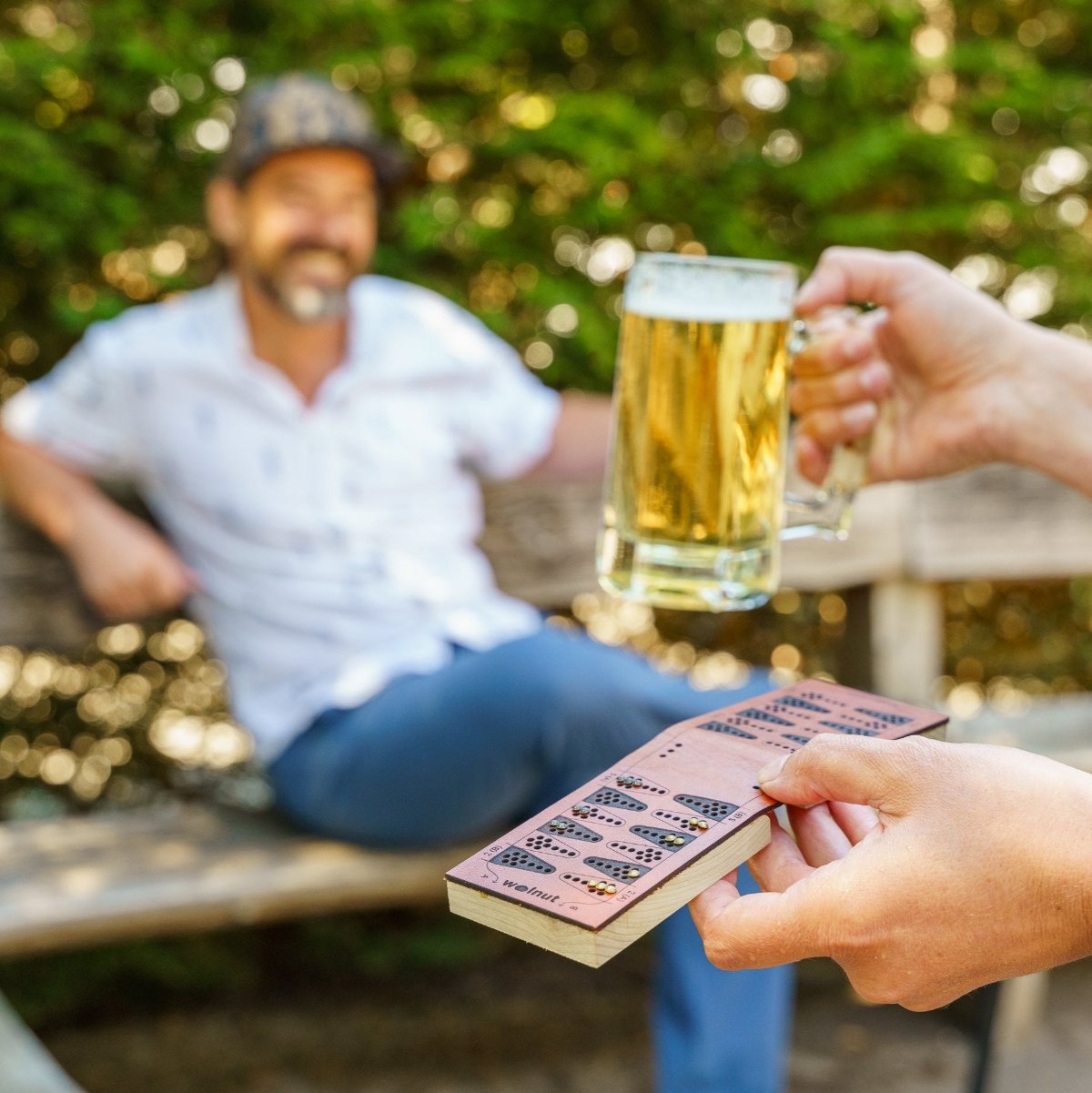 In the foreground, a standing person holds a beer in their right hand and a leather travel backgammon board with peg game pieces in their left hand, walking toward a man in the background who is blurred out but clearly relaxed and smiling, ready to play a game at the park or restaurant