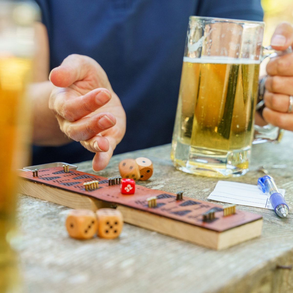 A person wearing a navy blue shirt is rolling wood dice on a picnic table next to a leather and wood travel backgammon board. They are rolling with their right hand and the dice are tumbling down from their hand inmotion, and in their left hand they&#39;re holding a stein of golden yellow beer. 