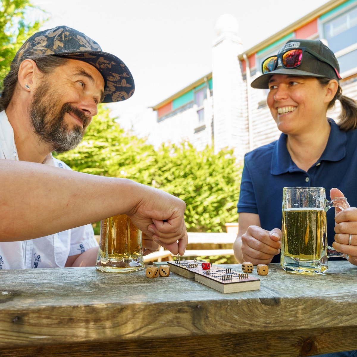 A man and a woman playing travel backgammon outdoors at a cafe holding beers and smiling at each other