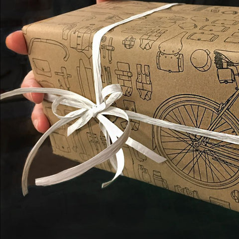 A hand holding a gift-wrapped present on a black studio background. The gift wrap is kraft paper with a black line drawing featuring a bicycle and tools and has a white paper raffia ribbon. All the materials are uncoated paper, recyclable and compost-able, as a green and eco-friendly choice. 