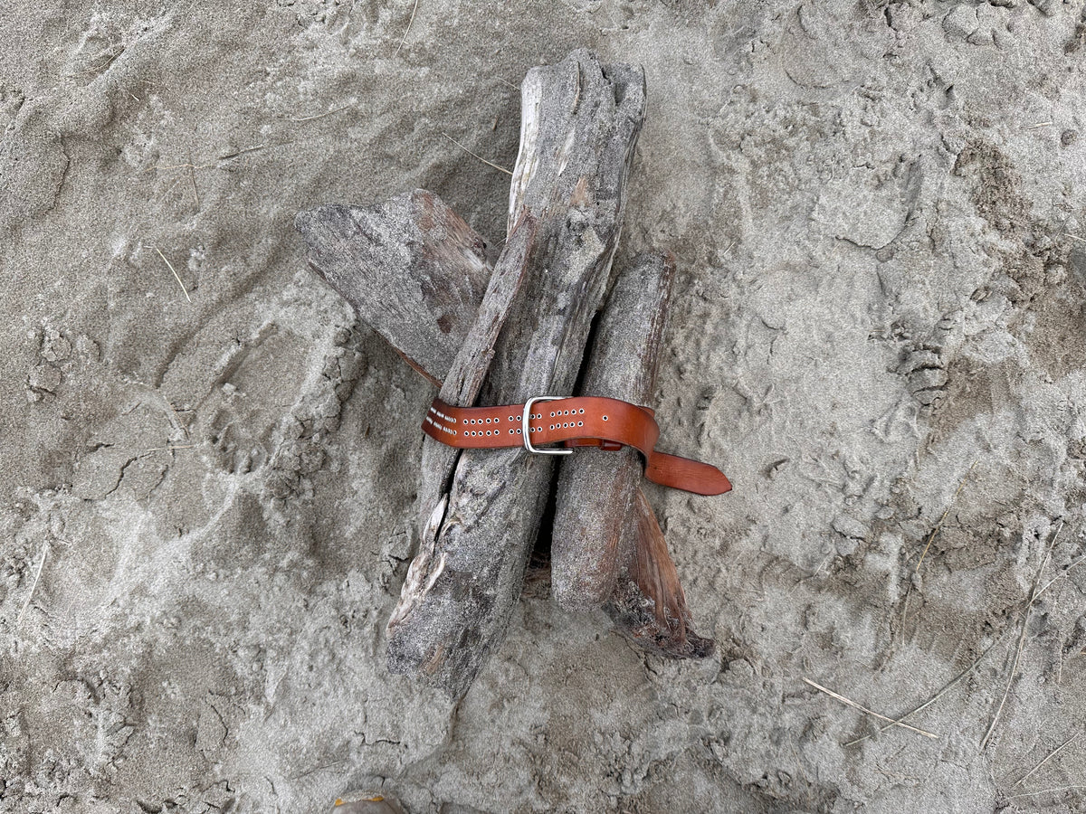 a bundle of gray driftwood logs gathered together and held in place with a tan leather cribbage board belt on a sandy beach