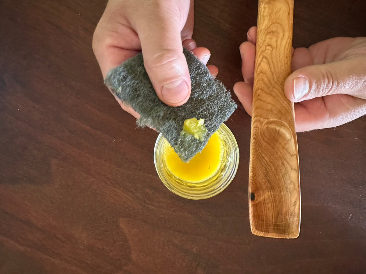 A person holding an abrasive pad with a dollop of yellow wood conditioner wax in one hand and a dry wood spoon in the other hand