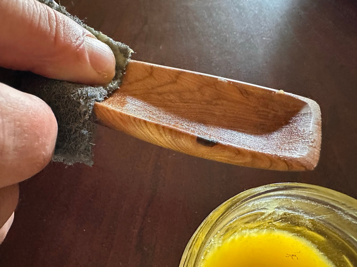 A person rubbing a wood spoon with  an abrasive pad, leaving the spoon shiny and wet-looking