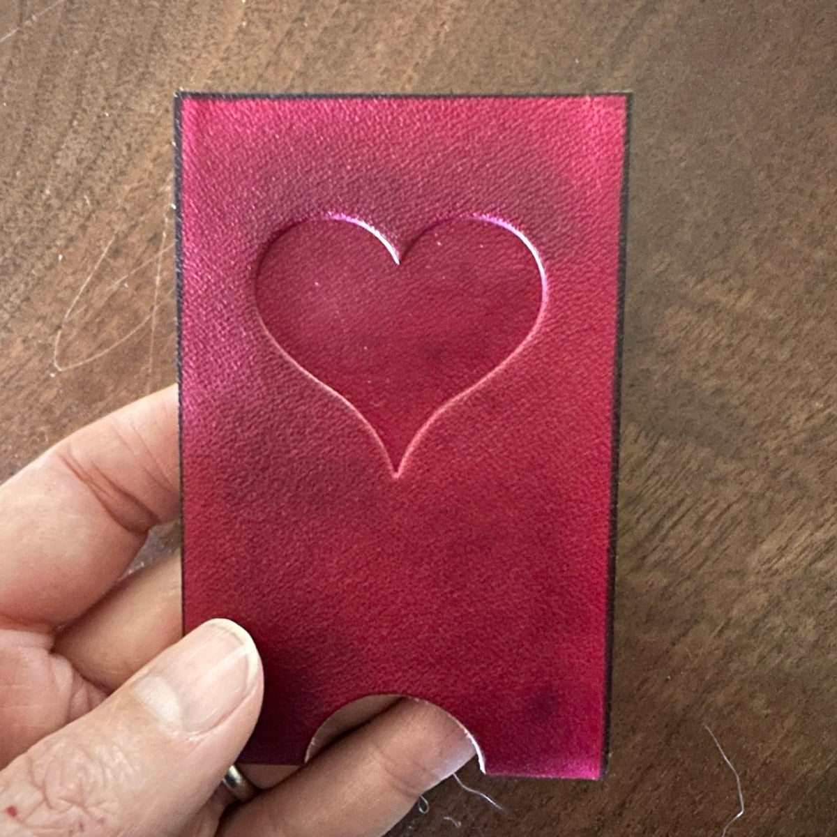 Hand holding a red leather card with a heart embossed in it over a wooden surface