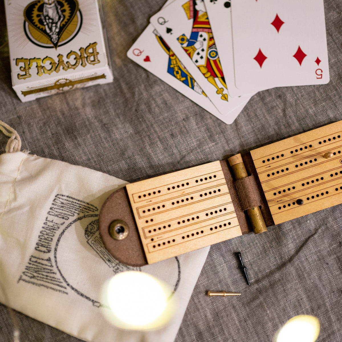 Open leather and wood travel cribbage board with cast metal person on top of a bedspread with a carrying pouch and a deck of cards, which is the &quot;Gift Set&quot;