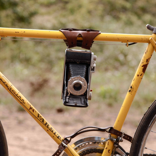 Vintage camera attached to a rusty yellow Schwinn bicycle frame with a brown leather cargo strap in front of a blurred natural background outdoors