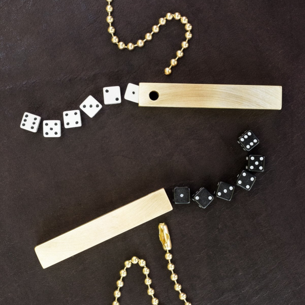 Two sets of travel dice on a dark brown leather table surface. Both sets show six dice coming out of a square brass tube. The dice are 7mm, smaller than standard. One has white dice and the other has black dice.