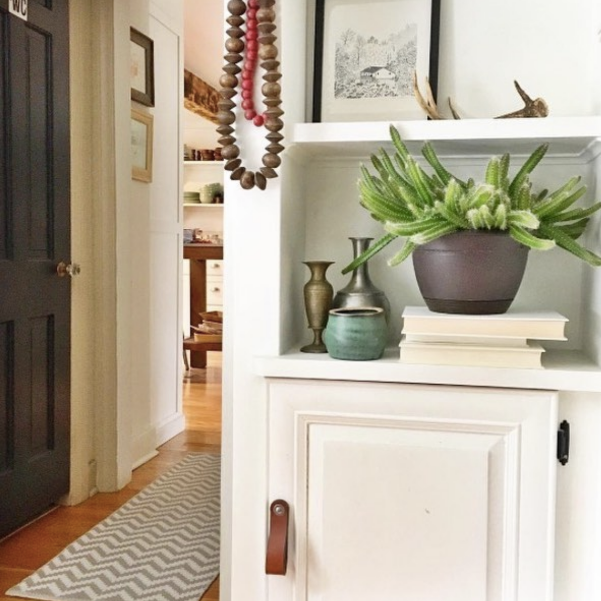 Customer photo of a creative artistic bohemian living room built-in shelf with potted plants and beaded necklaces and pretty turquoise vases. The cabinetry is white is there is a honey Hawthorne leather handle on the cabinet door. 