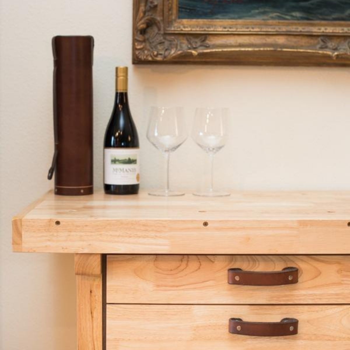 Blond hardwood side table with dark brown leather handles with a leather wine bottle case and two wine glasses. 