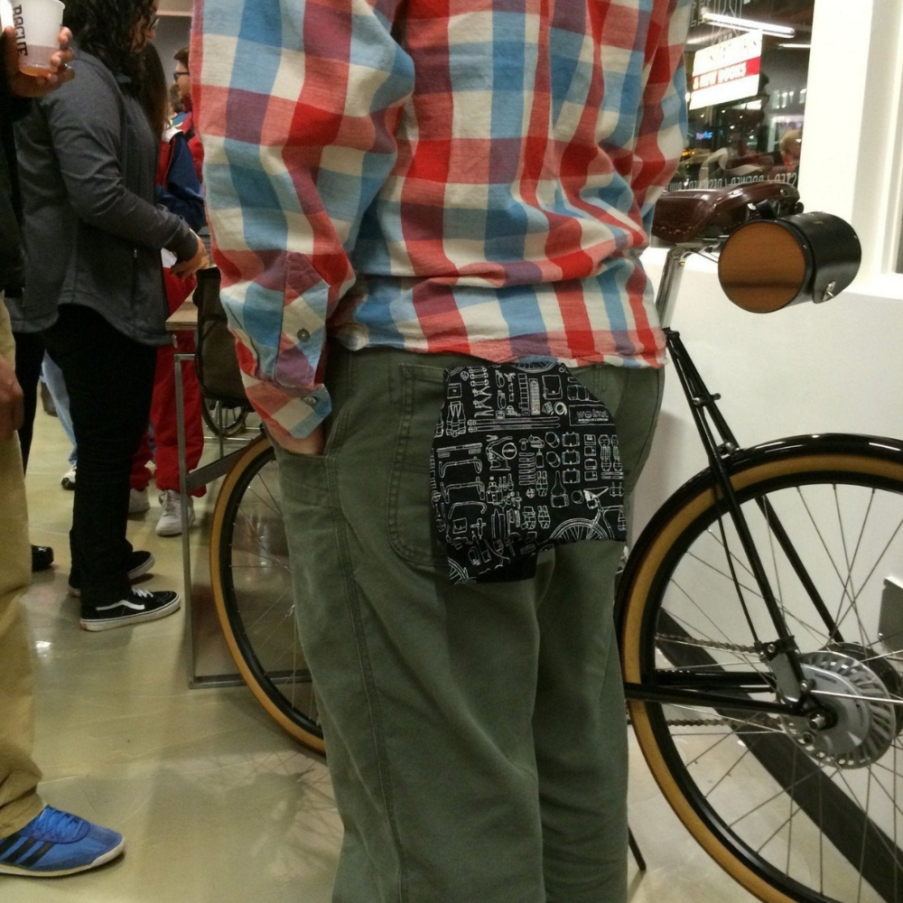 Black print bandana worn in left man&#39;s back pocket. The man is standing in a crowd holding his bike, which also carries the walnut studiolo black leather barrel bike bag.