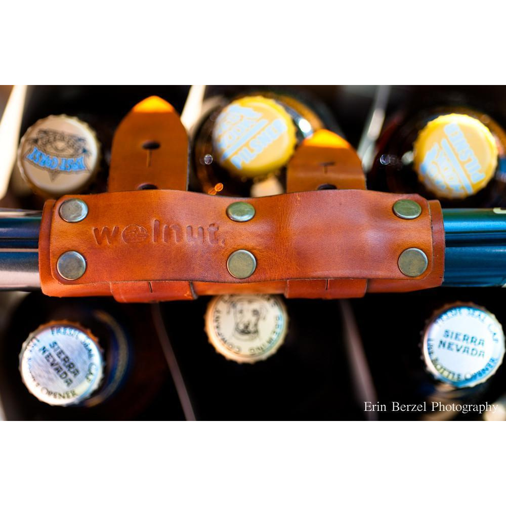 A close-up image of the honey leather variant of the 6-pack frame cinch. The beer carrier is attached to the top tube of a bike frame and is holding a yellow beer carton with six glass bottles. The cinch is embossed with the phrase walnut. Erin Bezel Photography watermark is in the corner.