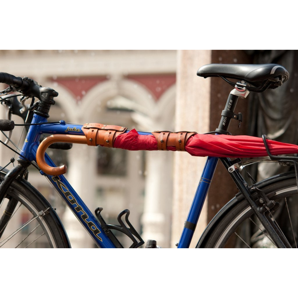 Two honey leather 6-pack frame cinch bicycle beer carriers are attached to the top tube of a blue bike. The bike beer holders are holding a red umbrella tight against the bicycle top tube.