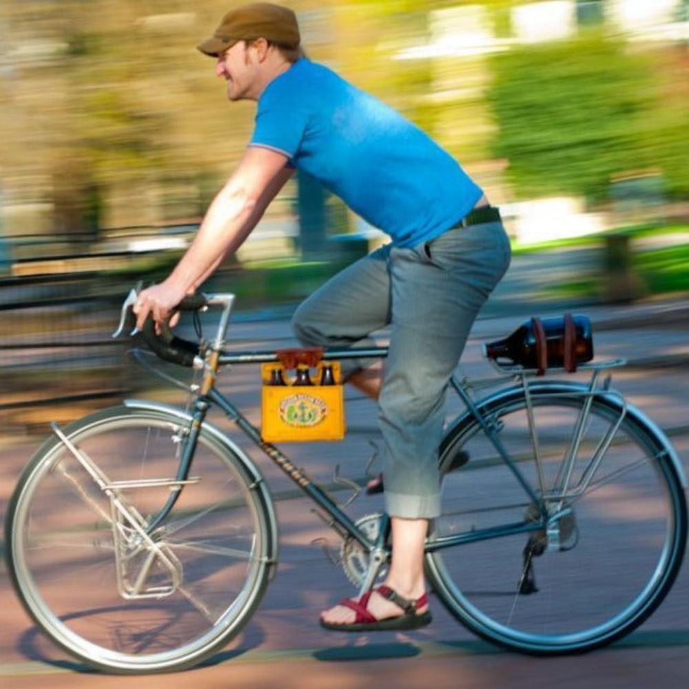 A man riding a bike with the background blurred. A yellow cardboard beer carton is attached to the top tube with the honey leather 6-pack frame cinch.