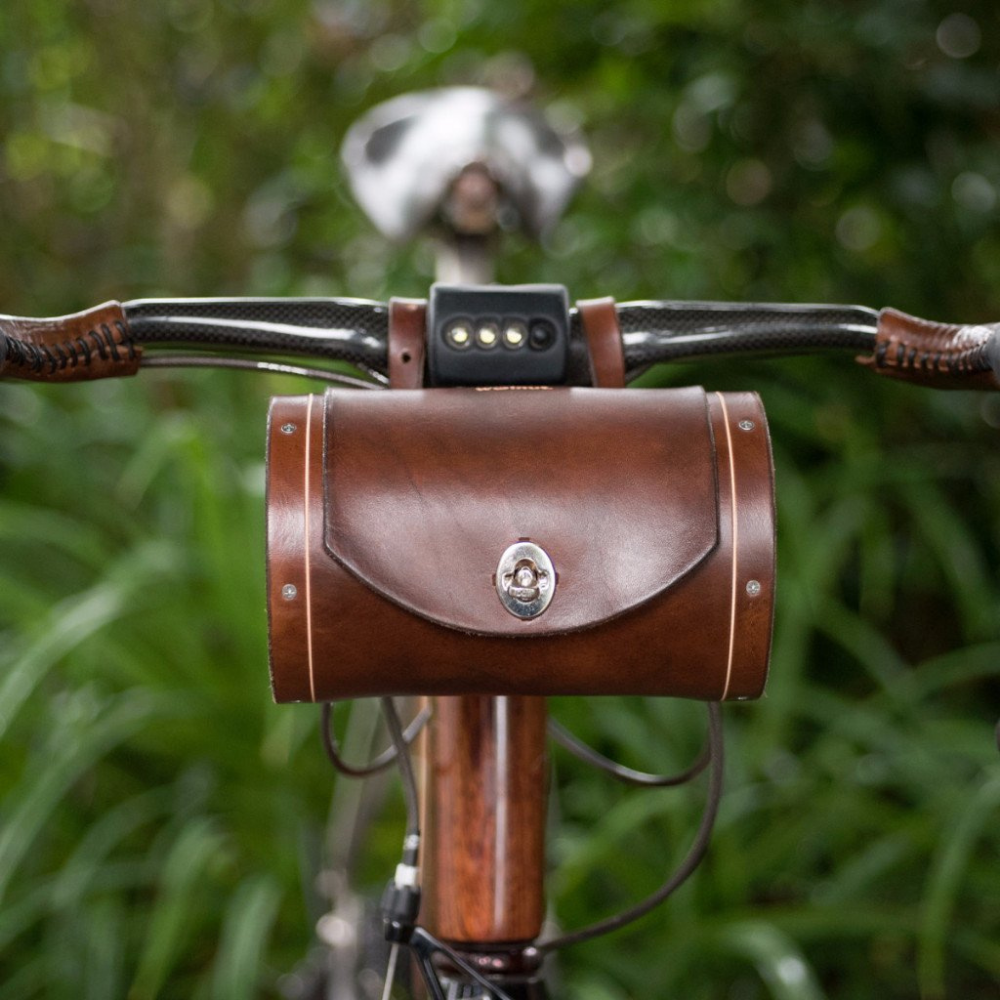 Dark brown leather bicycle barrel bag on handlebars with silver-look hardware and locking mechanism.