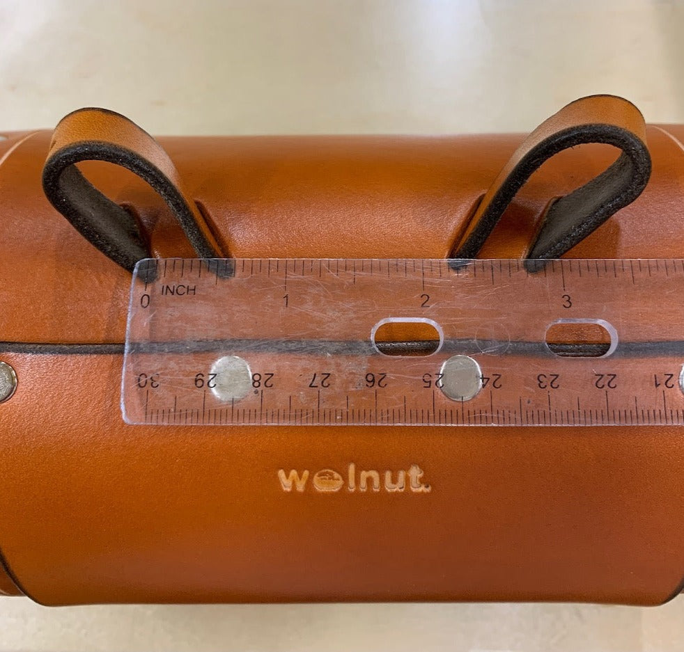 Honey leather variant of bicycle barrel bag being measured between two connecting points with clear ruler. The ruler shows that the difference between the two leather loops is three inches.