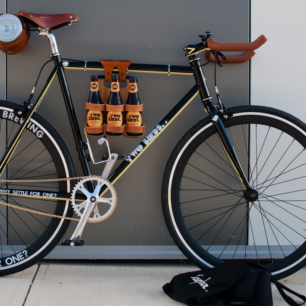 Honey leather and bamboo six-pack held on the top tube of a black bicycle with a 6-pack beer carrier, the 6-pack frame cinch on a custom branded bike for an Australian brewery