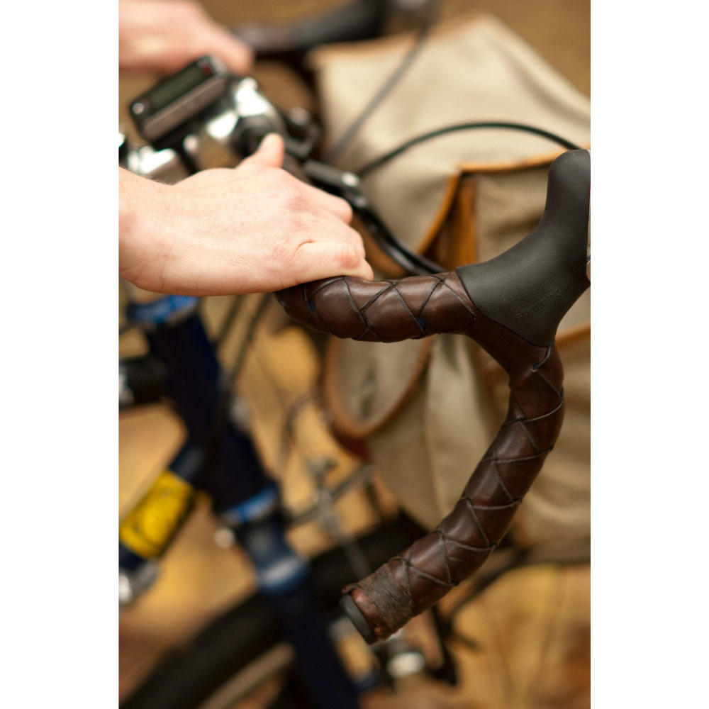 White hands on bicycle drop bar wrapped in dark brown leather bar wraps.